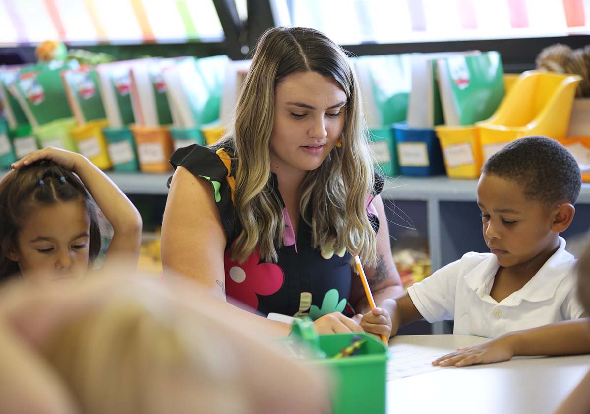HSA Teacher smiles while kneeling beside a young student in a classroom setting.